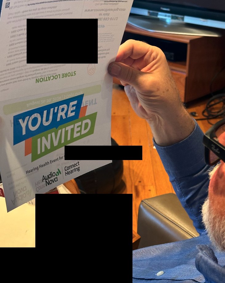 Seen from behind, a participant wearing Ray-Ban Meta glasses holds a piece of mail — a hearing-health event invitation — up to read it during a home visit; the name and address on the letter are blacked out.