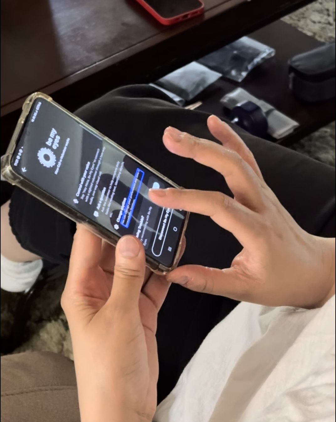 A participant's hands hold an Android phone showing the Be My Eyes onboarding screen, which explains that saying 'Hey Meta, Be My Eyes' starts a Call-a-Volunteer session from the glasses.