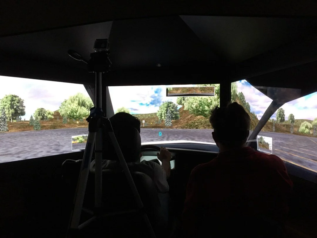 Inside the darkened driving simulator: a participant at the wheel and a researcher beside them face a wraparound projection of a rural road, with a GoPro on a tripod recording.