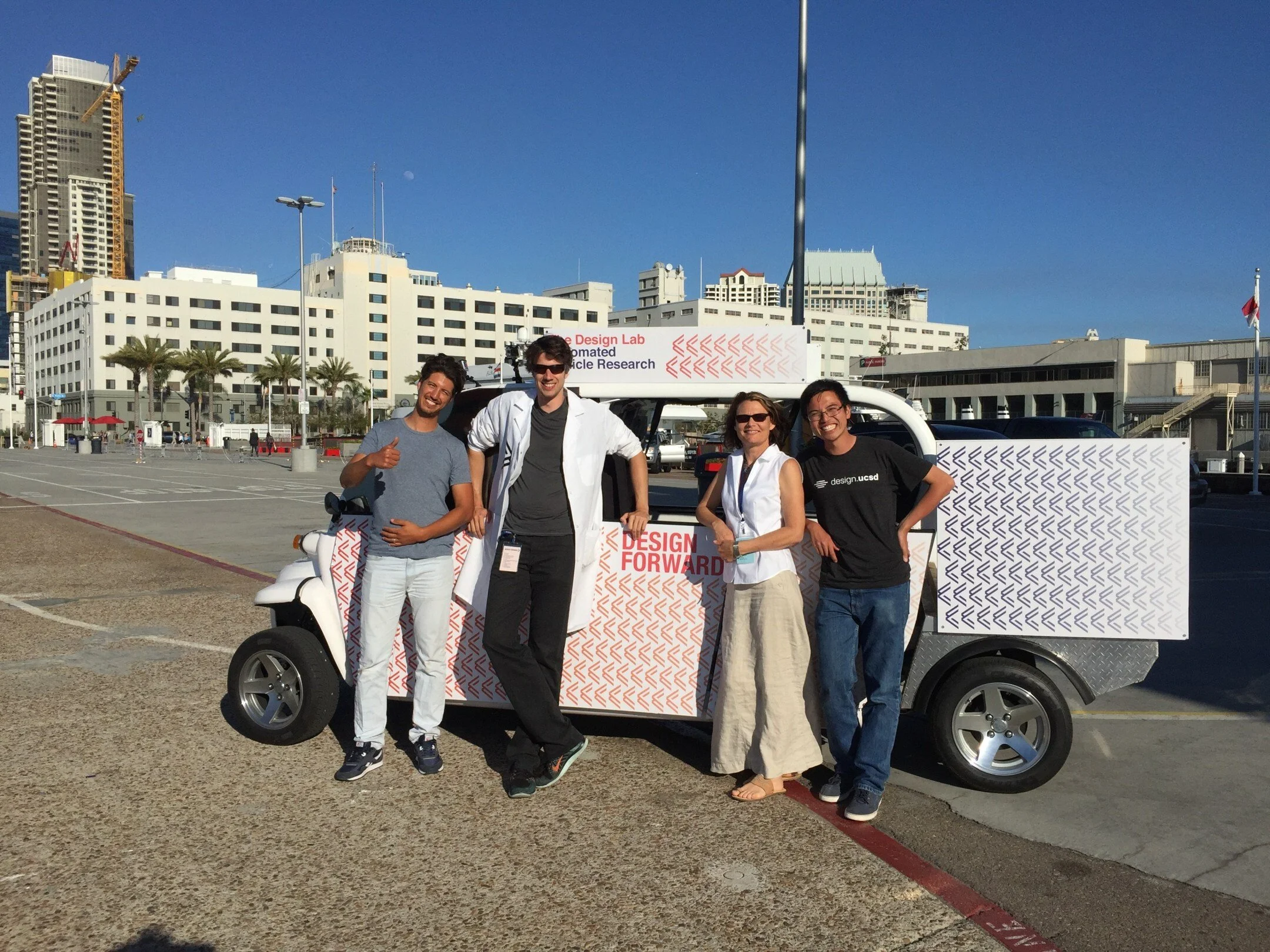 Four members of the Design Lab team, including Wesley in a design.ucsd shirt, pose beside the Design Forward golf cart on Broadway Pier.