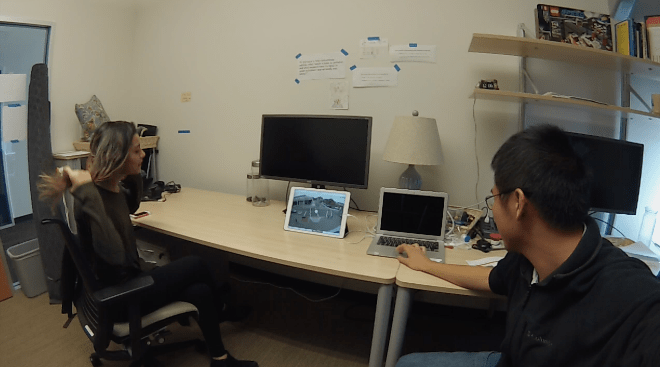 Wide-angle still of a lab interview: a participant gestures while watching traffic footage on a tablet, with the researcher seated across the desk.
