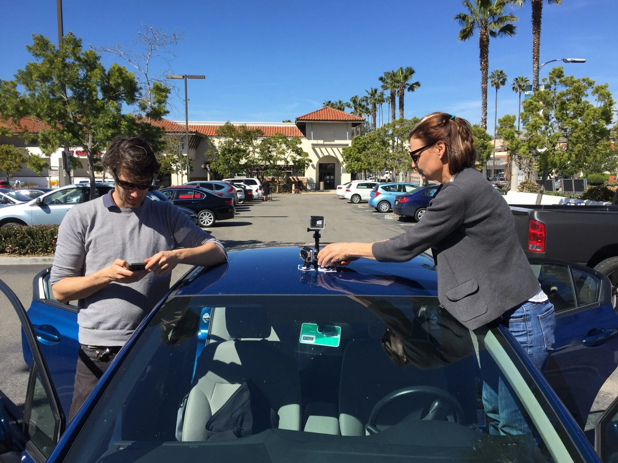 Malte and Colleen mount GoPro cameras to the roof of a blue sedan in a sunny parking lot before a seat-suit drive.