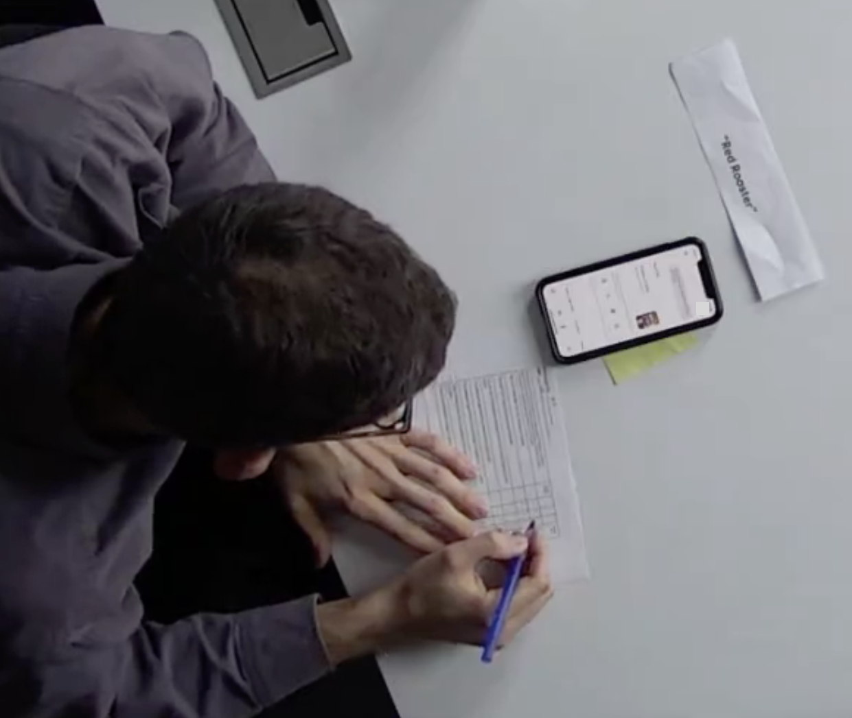 Overhead view of a lab session: a participant fills in a paper survey next to their own phone showing Assistant results, with a task card reading “Red Rooster.”