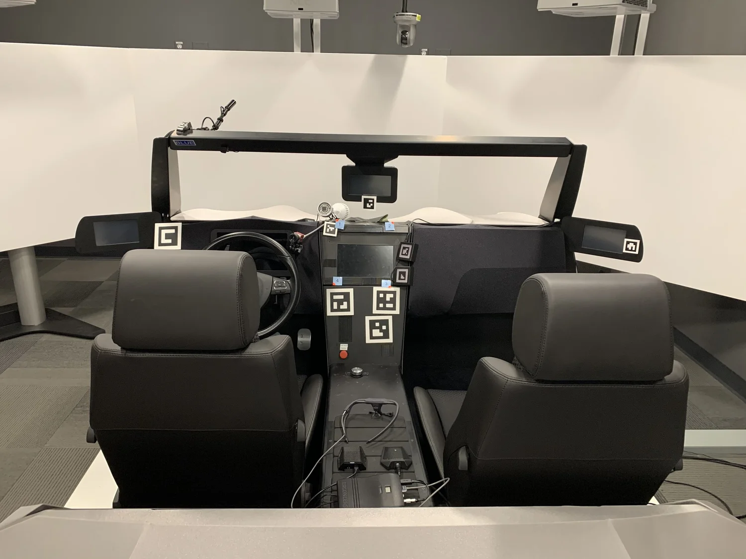 A driving-simulator rig: two car seats, a steering wheel, and a dashboard covered in fiducial markers, facing a wraparound projection screen.