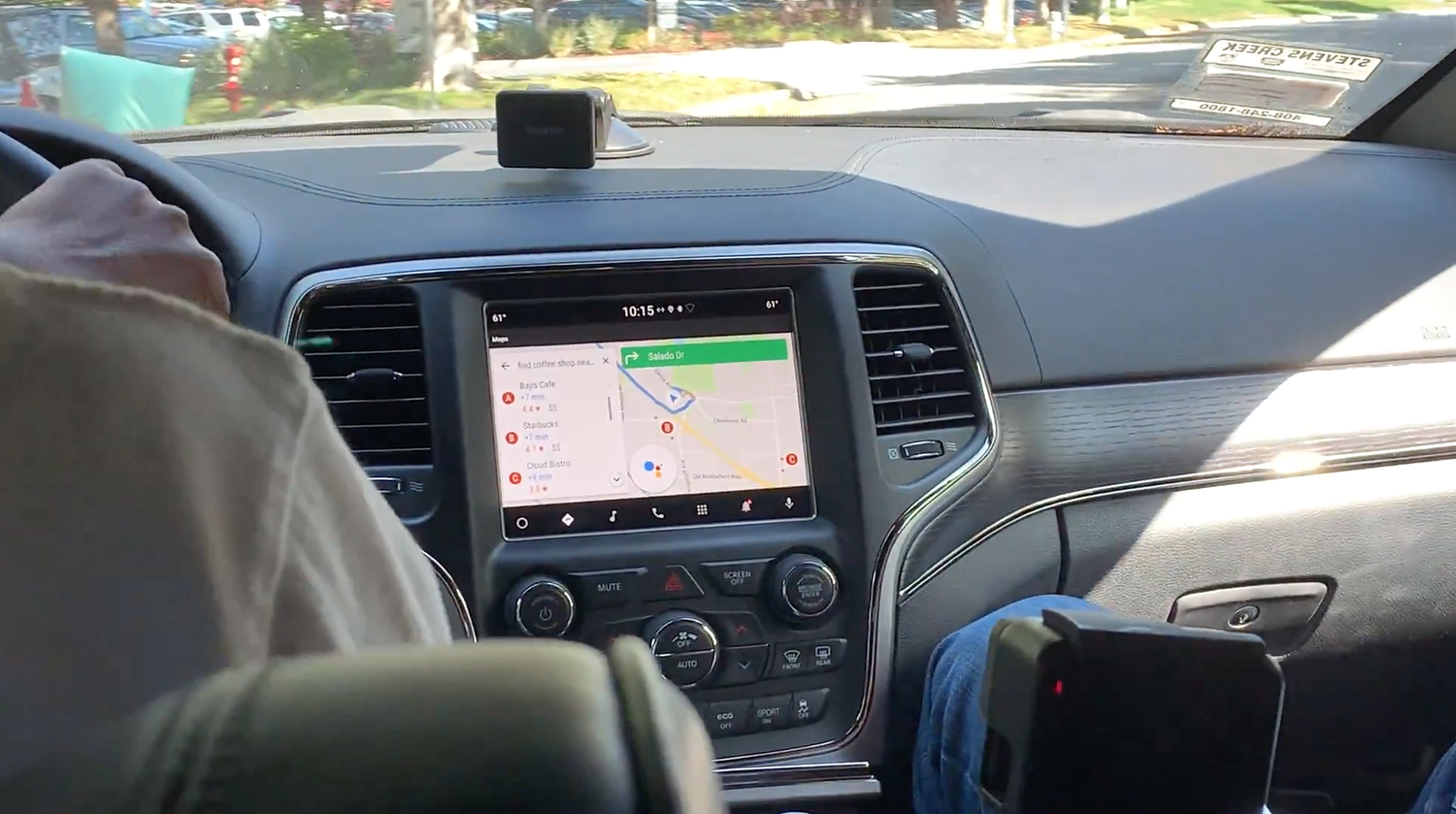 Over-the-shoulder view of a car's dashboard screen showing Google Maps with nearby coffee-shop results and the Assistant listening indicator, mid-drive.