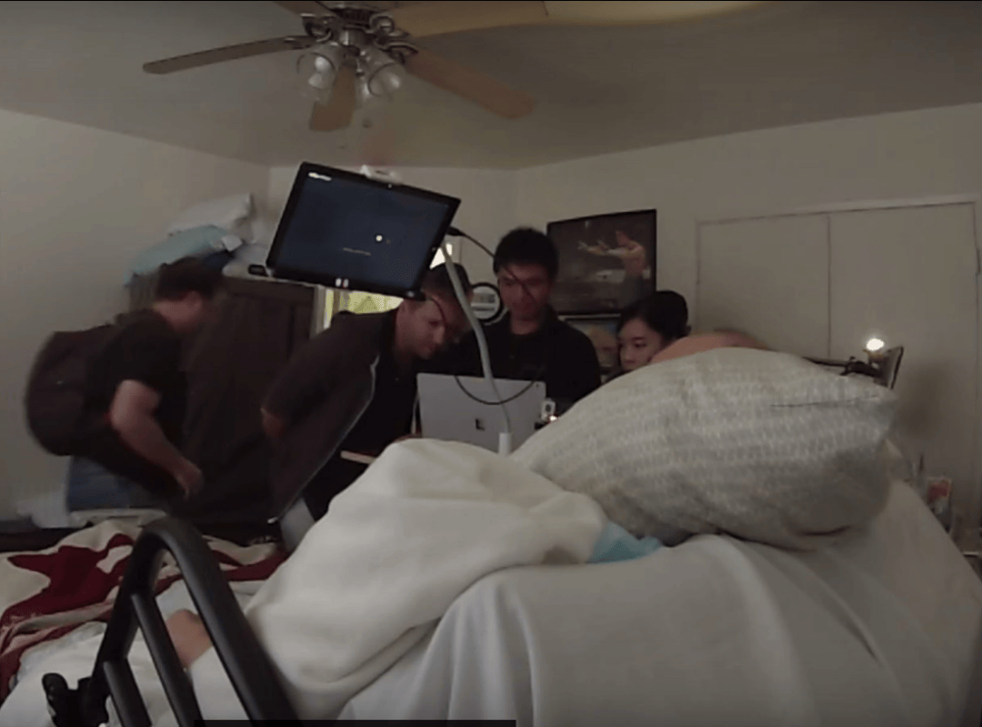 GoPro still of a home session: the team gathers around a Surface tablet on a gooseneck mount at a participant's bedside.