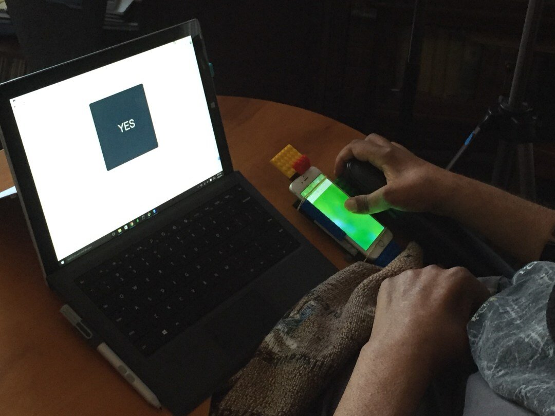 The prototype in use at a participant's home — a laptop screen shows a large YES tile while the participant's hand rests on a phone-based input rig mounted with LEGO bricks.