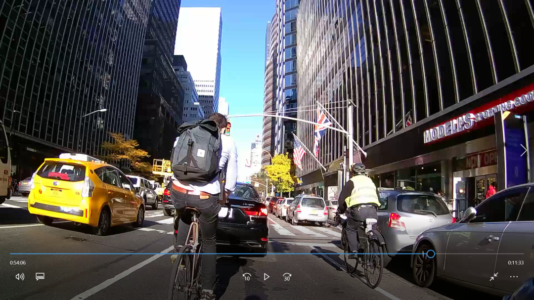 Handlebar-camera frame on a Midtown avenue: Brian rides ahead between a taxi and a black sedan, another cyclist to the right.