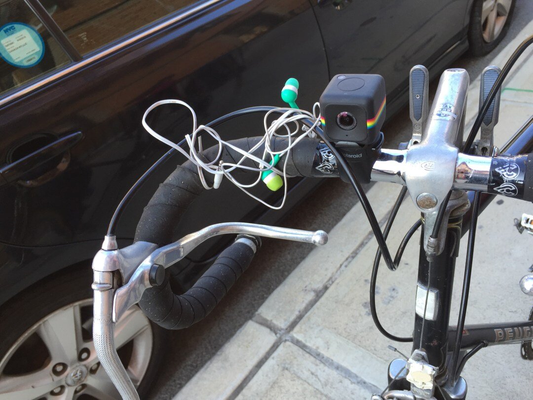 A small action camera mounted to the handlebars of a road bike with tangled earbuds beside it — the chase bike's recording rig.