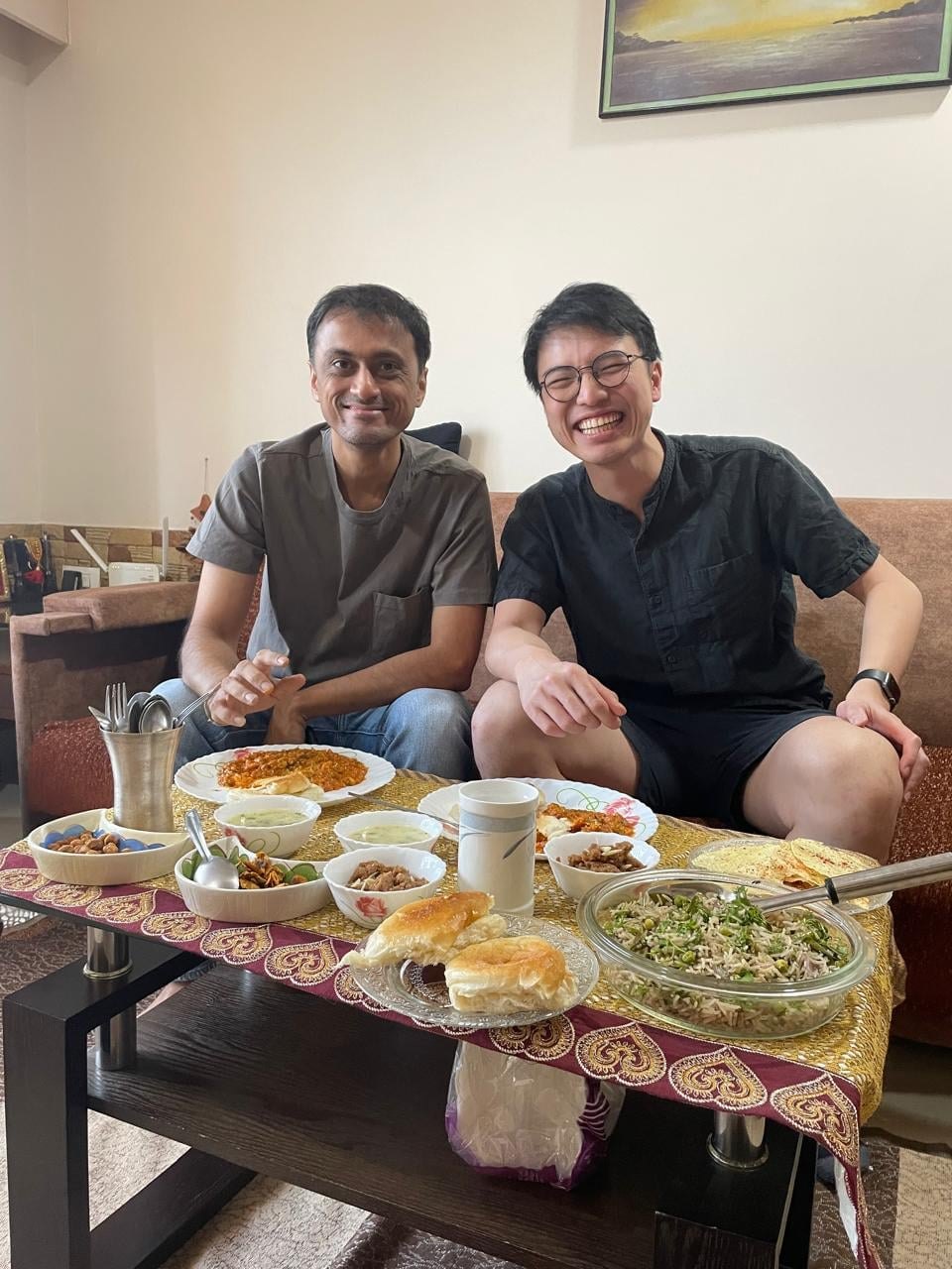 Two men sit side by side on a couch behind a low table set with lunch — pav bhaji, pulao, dal, papad, and small bowls of snacks on an embroidered maroon-and-gold cloth — both smiling at the camera.