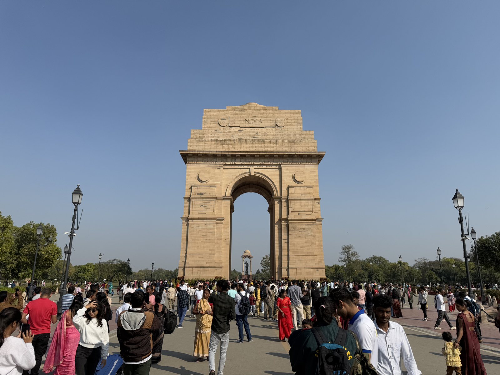 India Gate in Delhi on a clear day, crowds of visitors in the plaza beneath the arch, the canopy visible through it.