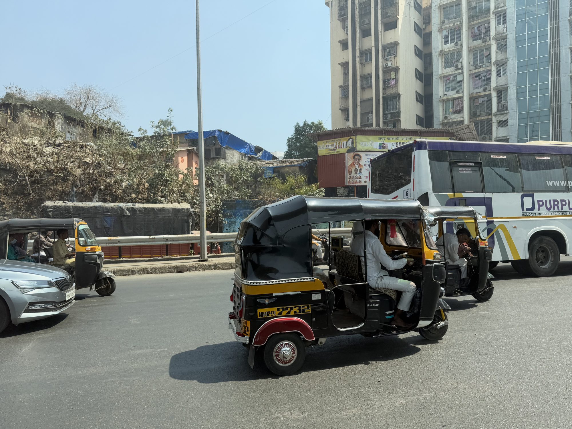 A black-and-yellow auto-rickshaw crosses a sunlit Mumbai road, a city bus and high-rise apartment blocks behind it.