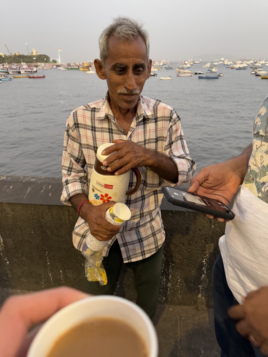 A chai vendor on the Mumbai seafront pours from a flask while a customer holds up a phone to pay by QR code; a cup of chai in the foreground, fishing boats behind.