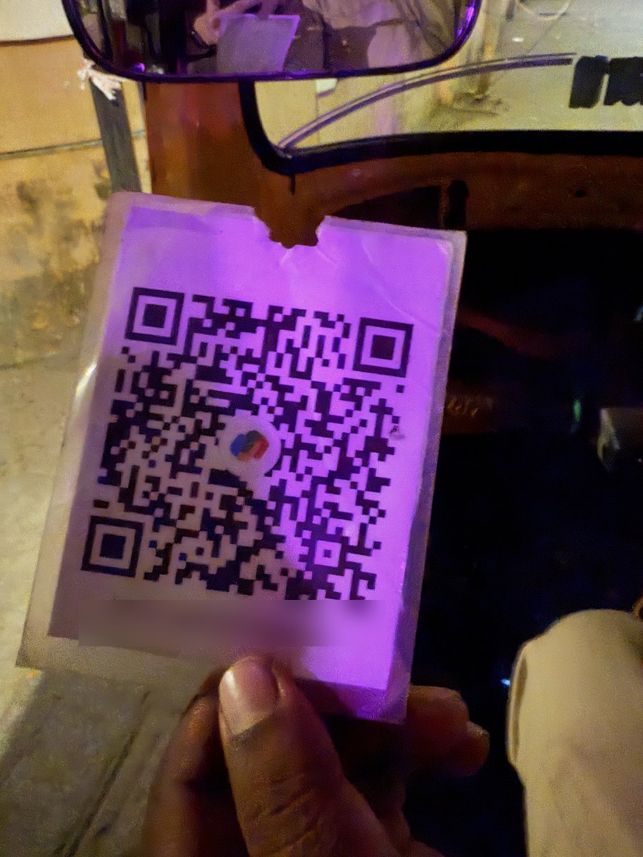 A laminated UPI QR code card held up inside an auto-rickshaw at night, lit purple by the cabin light.