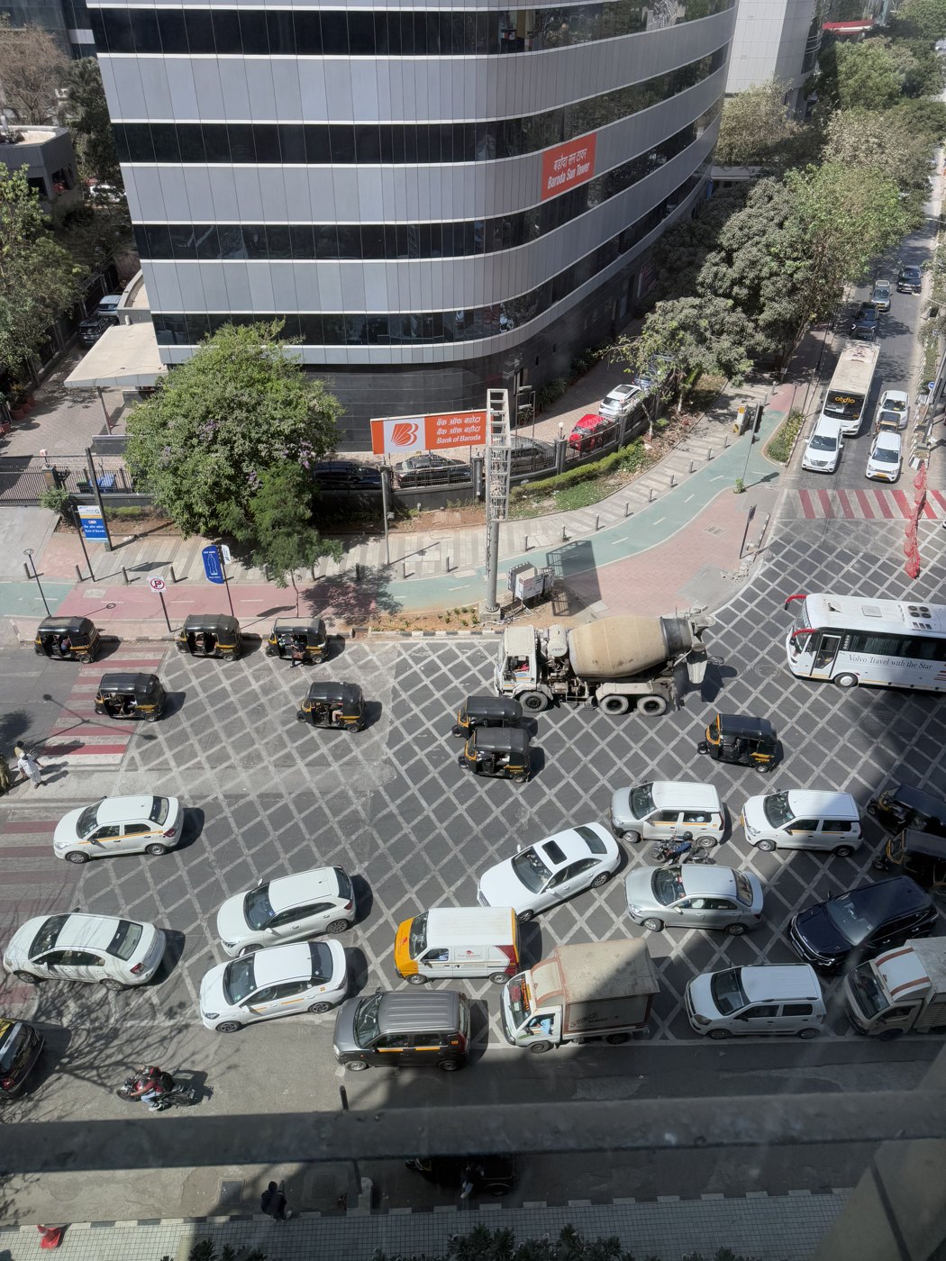 A Mumbai intersection seen from high in an office tower: cars, black-and-yellow taxis and auto-rickshaws, a cement mixer, and a coach all crowd a hatched junction box in front of the Bank of Baroda tower.