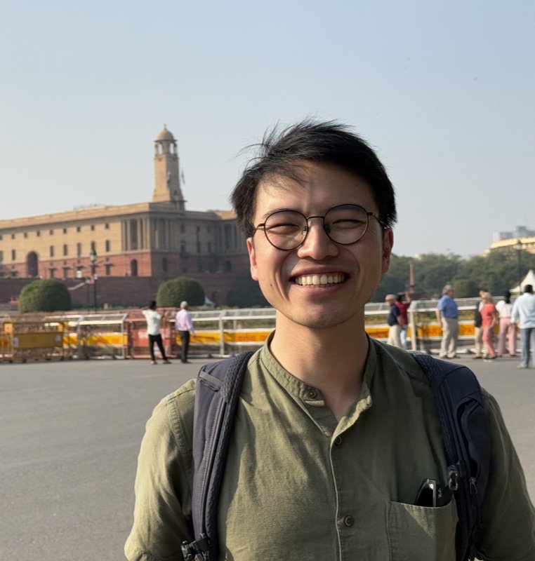Wesley Chan smiling in round glasses and an olive shirt, the Secretariat Building in New Delhi behind him.
